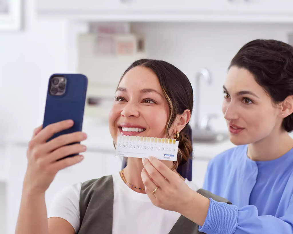 A woman smiles while taking a selfie with a smartphone. Another woman holds a teeth color chart beside her, indicating a dental check in a bright clinic.